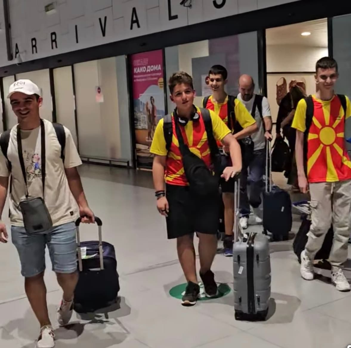 Натпреварувачите со куфери пристигнуваат на аеродром под натпис „Arrivals“.