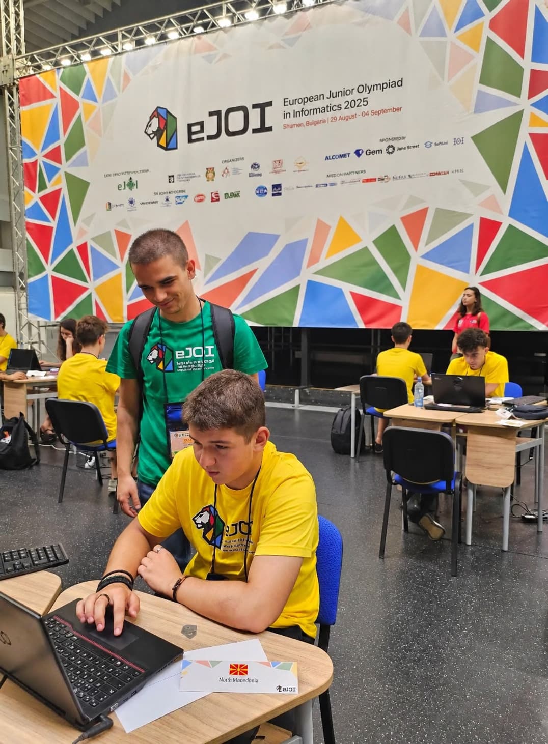 Натпреварувач работи на лаптоп пред банер на EJOI, Европската јуниорска олимпијада по информатика.